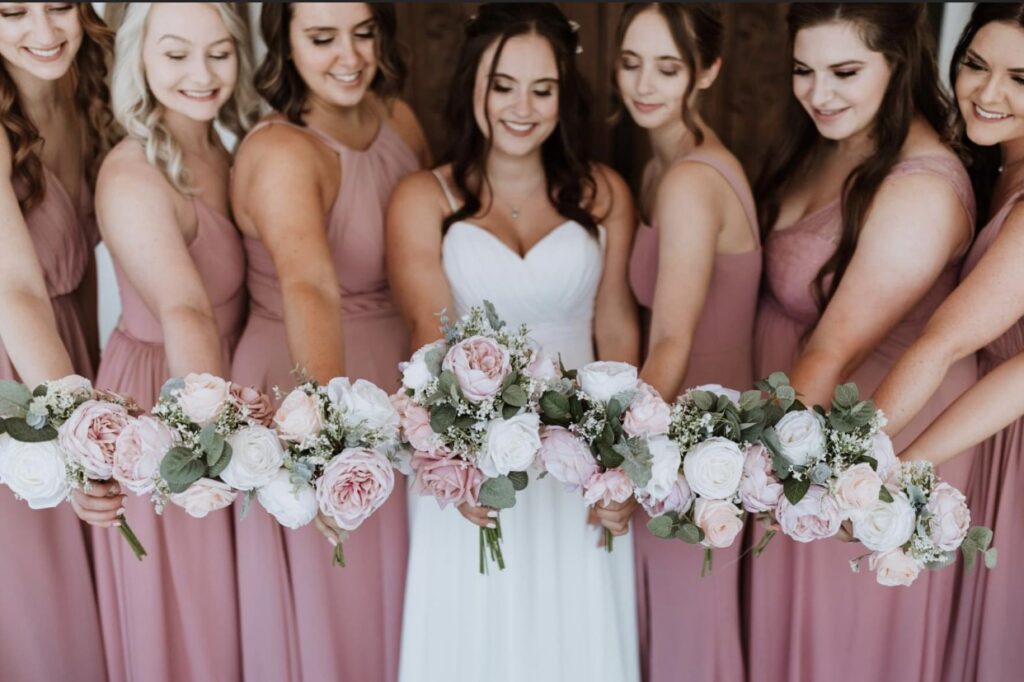 The bride and her bridesmaids who are dressed in blush gowns hold out their pink and white bouquets.