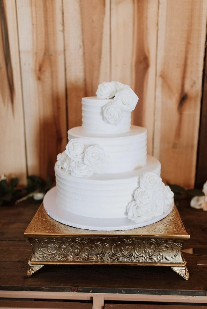 An all white wedding cake from Julia's Homestyle Bakery. It is decorated with with white roses and sits on a square gold cake stand. 