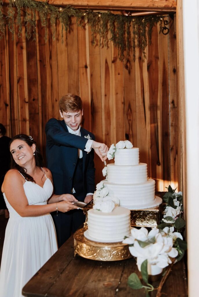 The bride and groom cut their all white wedding cake from Julia's Homestyle Bakery as they smile at their guests. It is decorated with with white roses. 