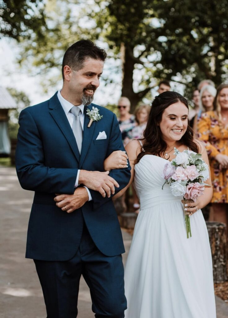 The father of the groom, dressed in navy, excitedly walks the bride down the aisle as she holds a white and blush bouquet of flowers.