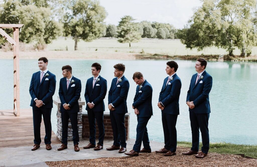 Groomsmen dressed in navy suits  stand lakeside at a wedding at Steel Magnolia Barn. 