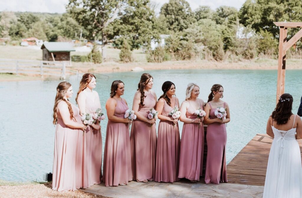 Ombre bridesmaids dressed in shades of rose and blush stand lakeside at a wedding at Steel Magnolia Barn. 