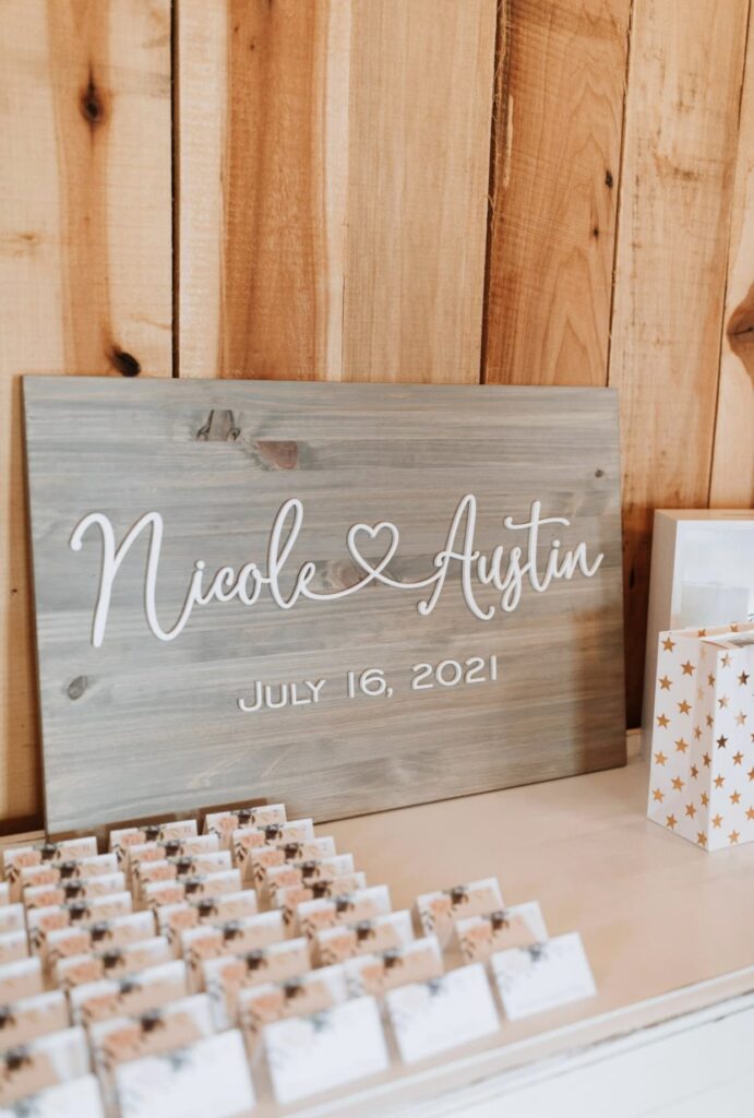 A barn wood sign with the bride and groom's names sits on a white table with seating cards and gifts. 