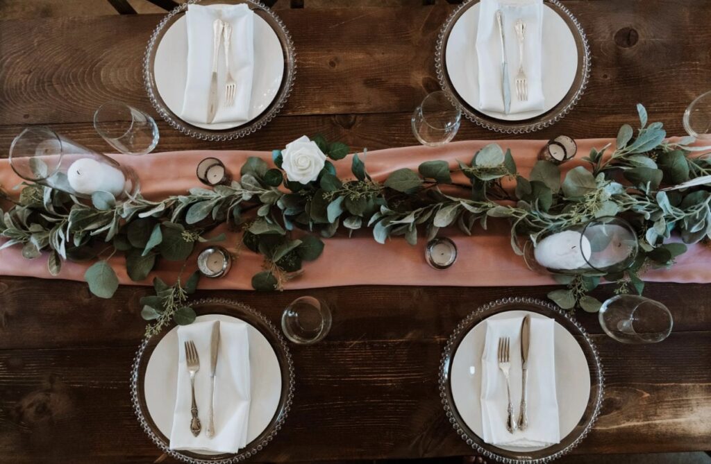Glass beaded chargers with ivory plates, white napkins and gold flatware sits on a farm table decorated with a rose colored table runner, a garland of greenery accented with white roses and candles. 