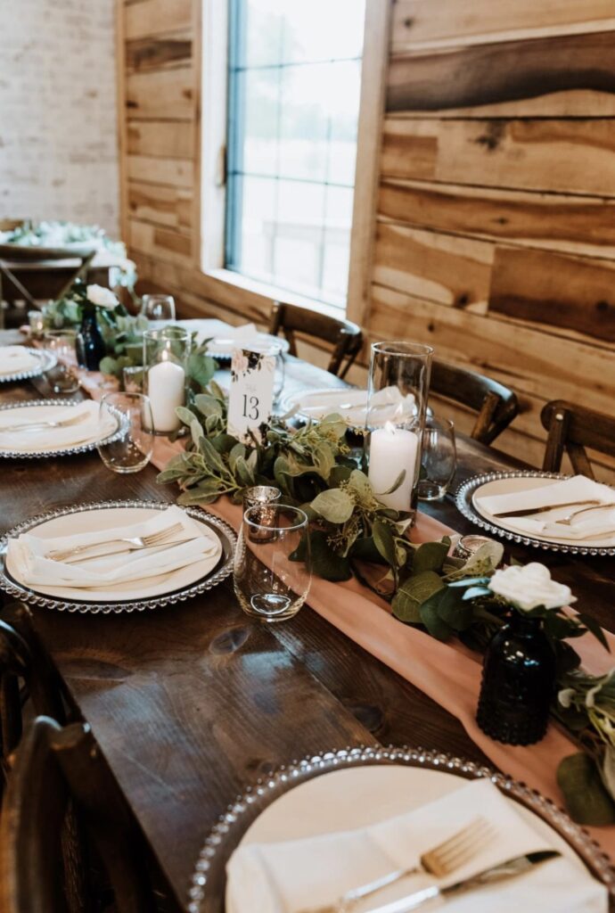 A farm table decorated with glass beaded chargers with ivory plates, white napkins and gold flatware accented with a rose colored table runner with a garland of greenery accented with white roses and candles. 