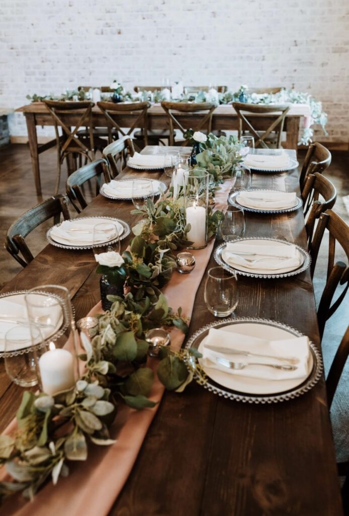 A long farm table with glass beaded chargers with an ivory plates and gold flatware is decorated with a garland of greenery and candles on top of a rose colored table runner. 