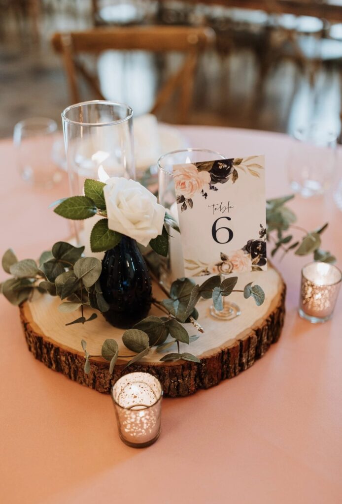 A wood slice with candles, greenery and white roses sits on top of a rose colored table cloth 