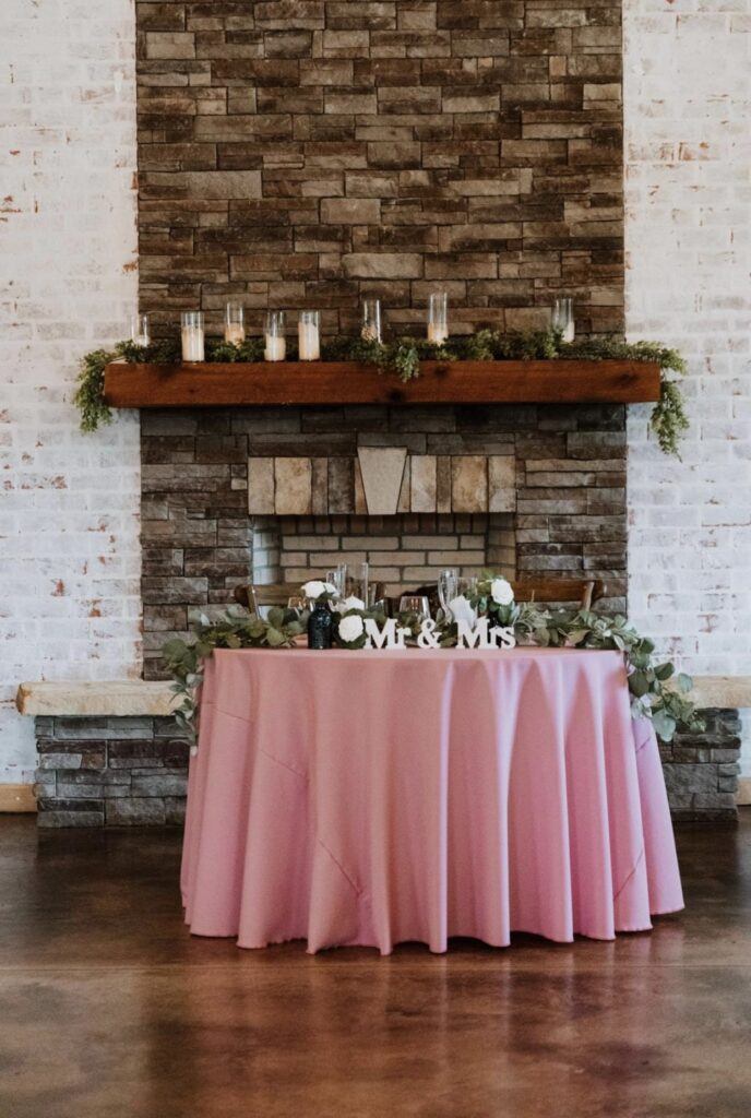 The stone fireplace is the backdrop of the bride and groom's rose colored sweetheart table which features candles, greenery and a Mr & Mrs sign. 