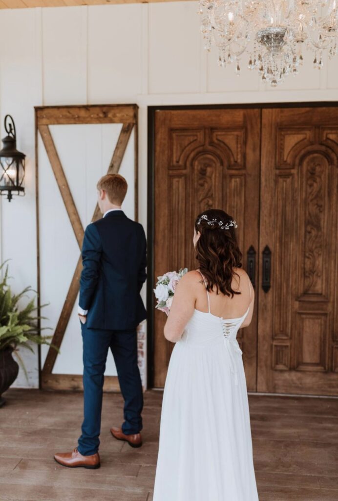 The bride approaches the groom for their first look in front of large wooden doors while holding her bouquet. 