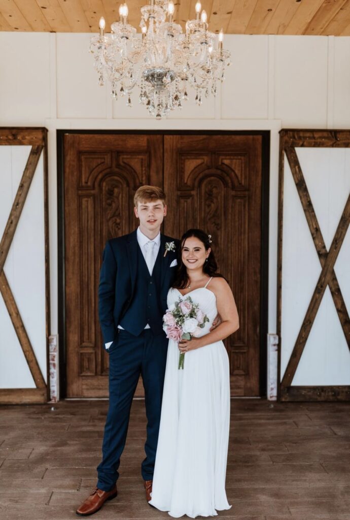 The bride holding her pink bouquet, stands next to the groom who is wearing a navy suit in front of the dark wood doors of Steel Magnolia Barn. 