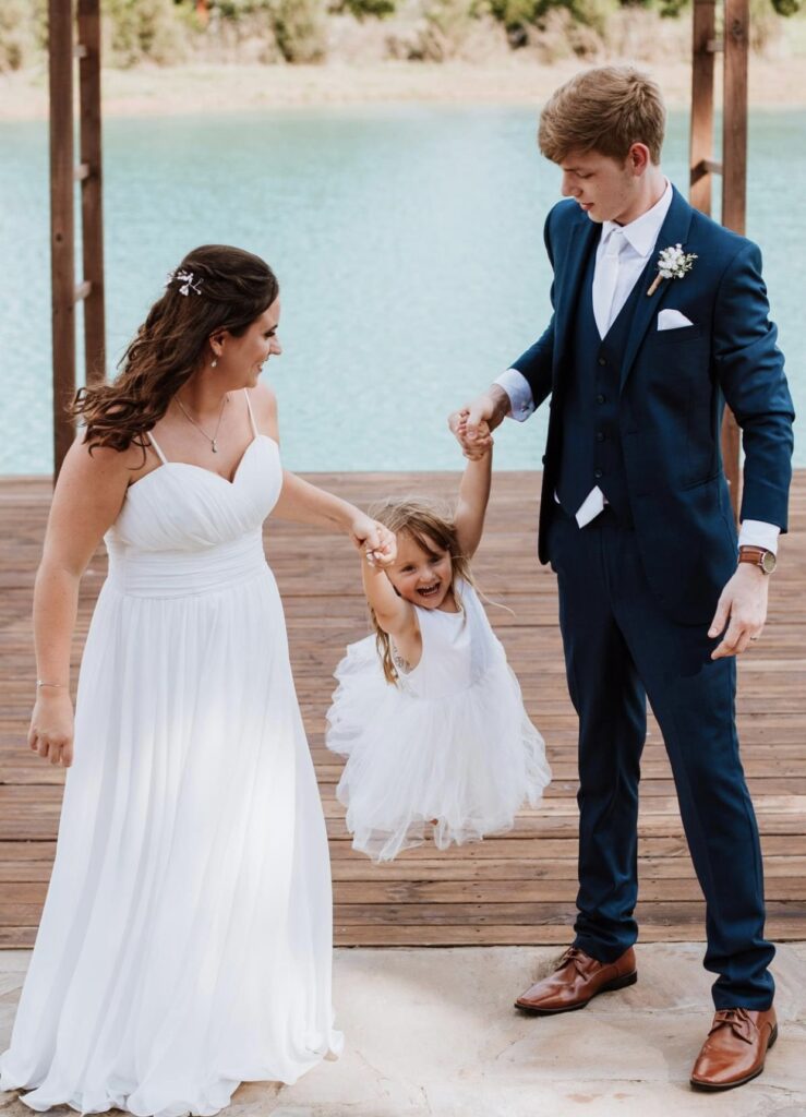 Bride and groom stand on the lakeside dock as they swing the flower girl in the air.