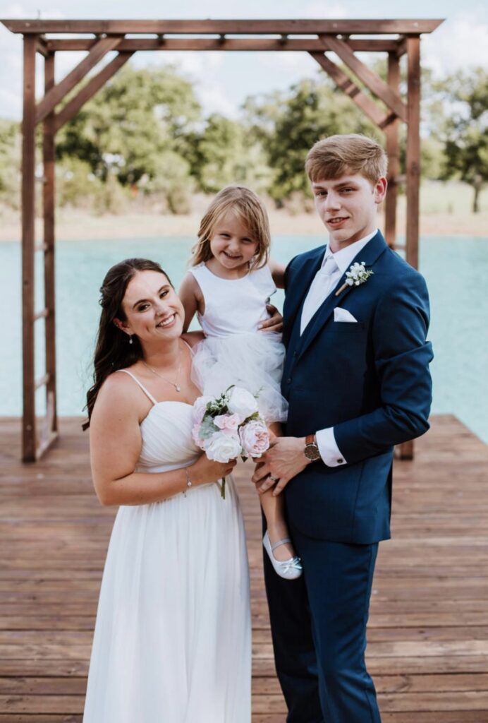 Bride and groom stand on the lakeside dock smiling as they pose with their daughter, the flower girl.