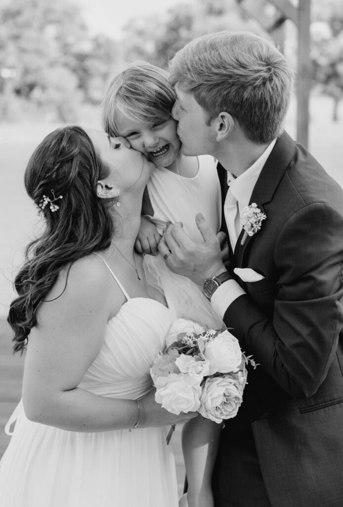 The bride and groom both lean in to give the flower girl kisses on her cheeks as she giggles at all the attention. 