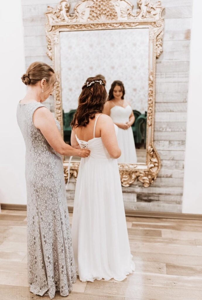 The mother of the bride, dressed in silver, helps the bride fasten the back of her dress closed and she looks on in a large gold framed mirror.