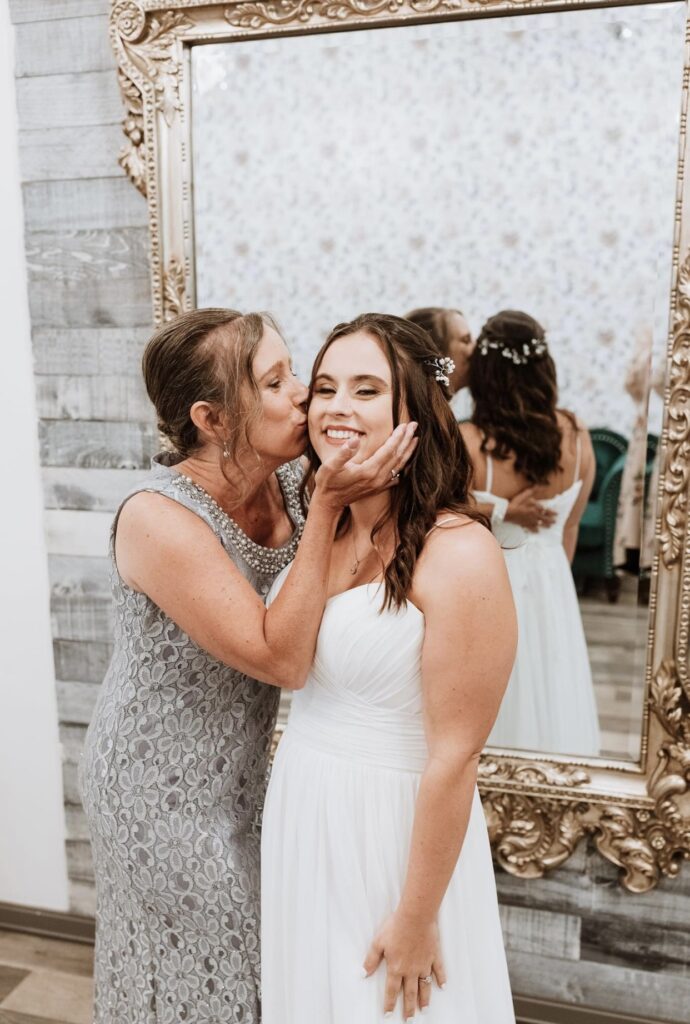 The mother of the bride, dressed in silver, gives her daughter a big kiss on the cheek.