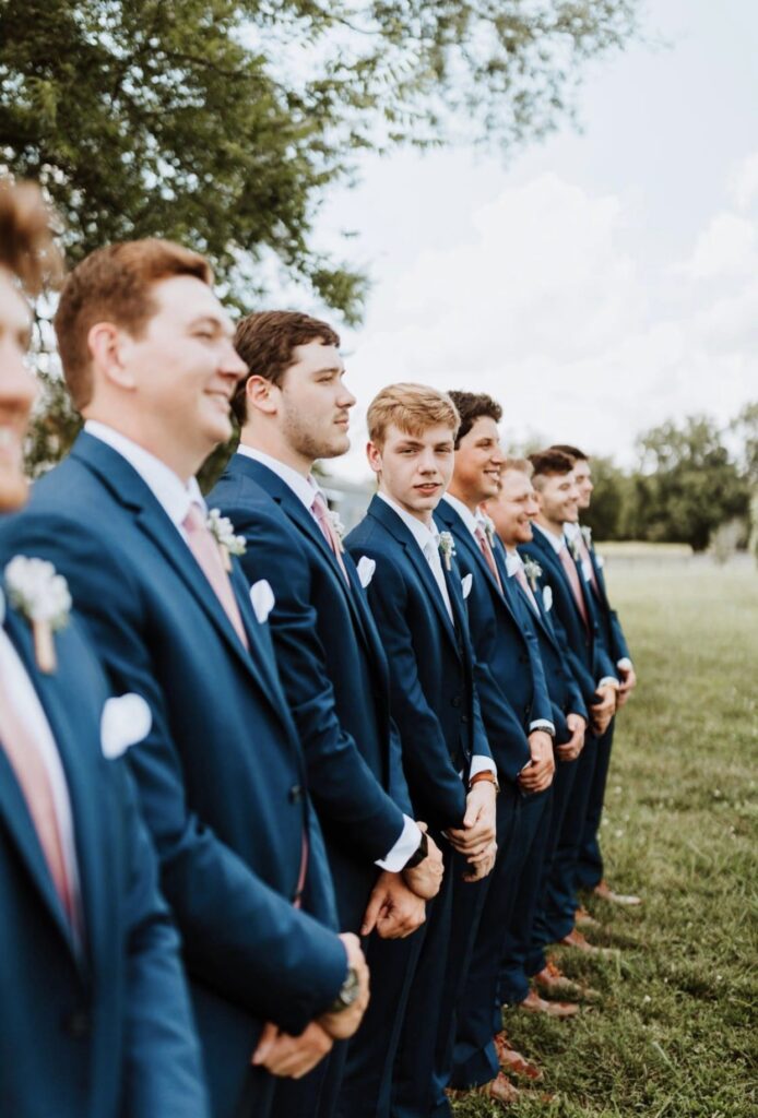 The groom lines up with his groomsmen in a lush green field. They are dressed in navy suits with blush ties and pocket squares. 