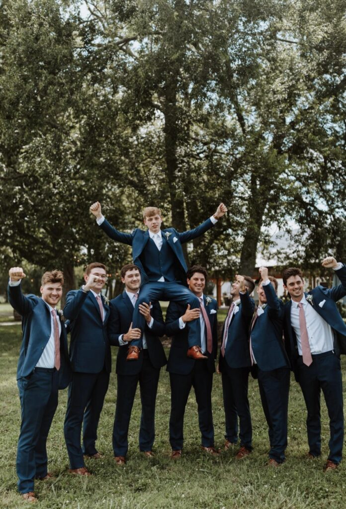The groom is lifted up in celebration by his groomsmen. They are dressed in navy suits with blush ties and pocket squares. 