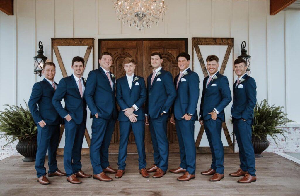 The groom lines up with his groomsmen in front of the wooden doors of Steel Magnolia Barn. They are dressed in navy suits with blush ties and pocket squares. 