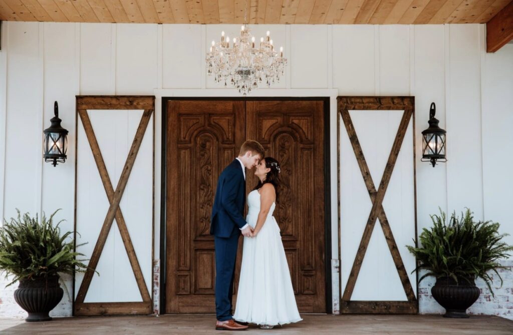 The bride and groom touch foreheads in front of the dark wood front doors of Steel Magnolia Barn. A crystal chandelier is suspended above them. 