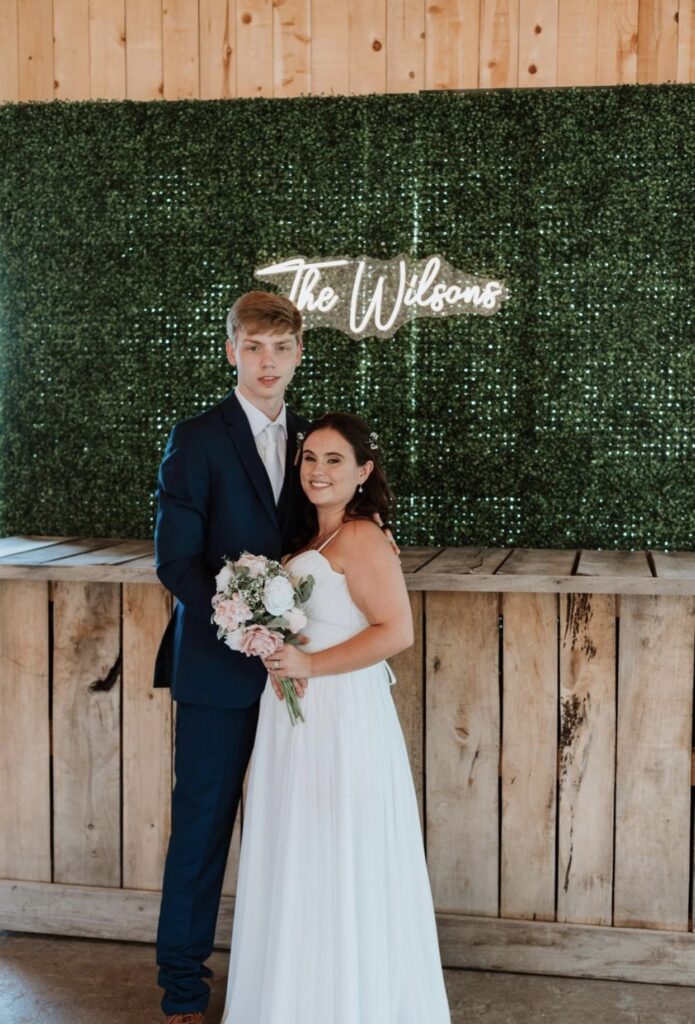 The bride and groom stand in front of a boxwood wall with a neon sign behind them declaring their new last name. The bride is holding a white and blush bouquet.