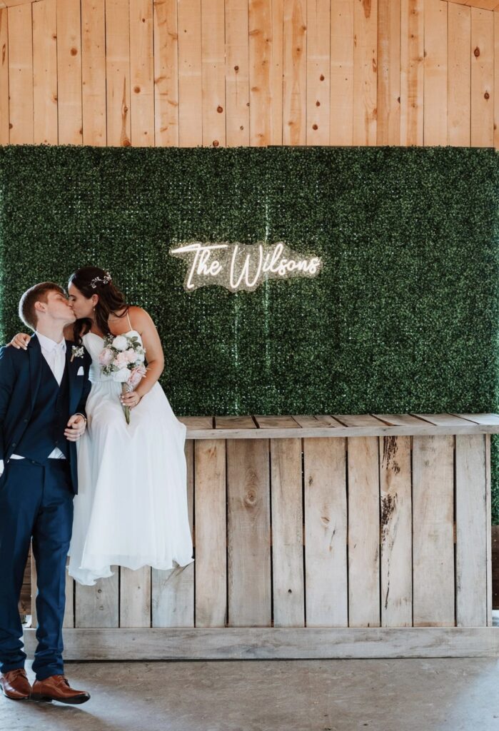 The bride and groom kiss in front of a boxwood wall with a neon sign behind them declaring their new last name. The bride is holding a white and blush bouquet.