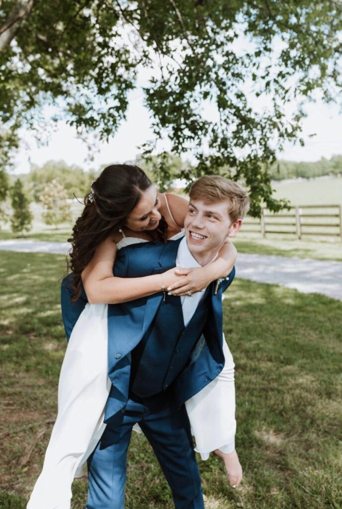 The groom, in a blue suit gives the bride a piggy back ride as he laughs and smiles up at her. 