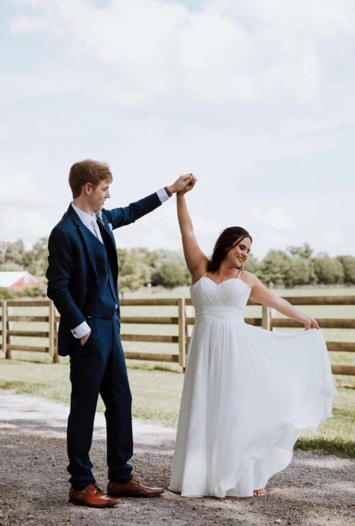 The groom, wearing a blue suit, spins the barefoot bride in a green field.