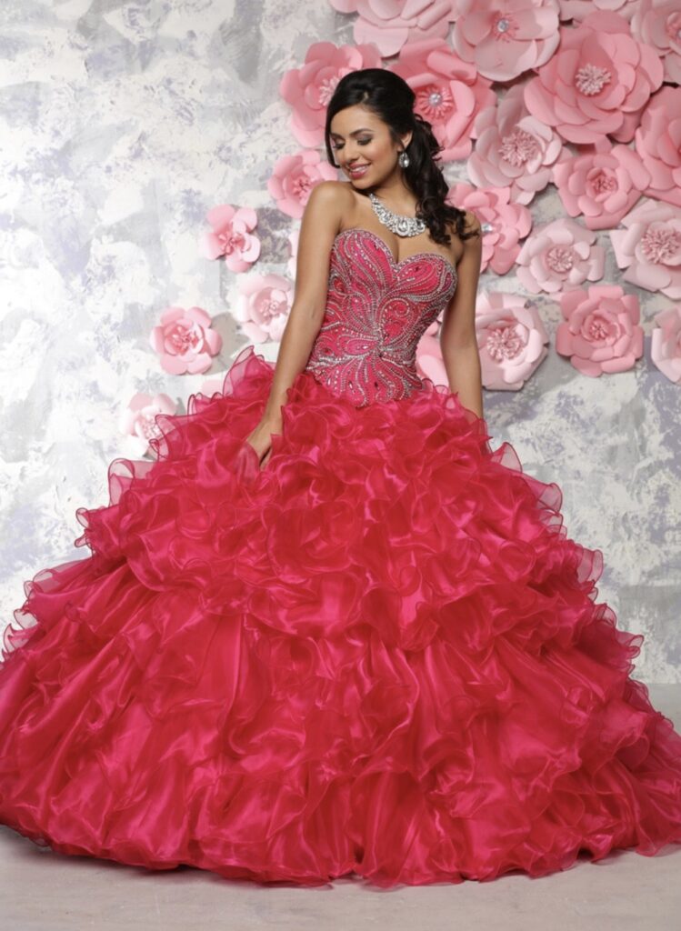 Quinceanera wearing a hot pink and silver ballgown with a ruffled skirt in front of a large paper flower wall.
