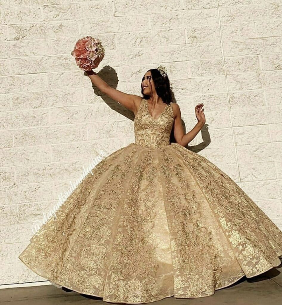 Quinceanera wearing a gold ballgown and tiara holding a gold and pink paper flower bouquet.