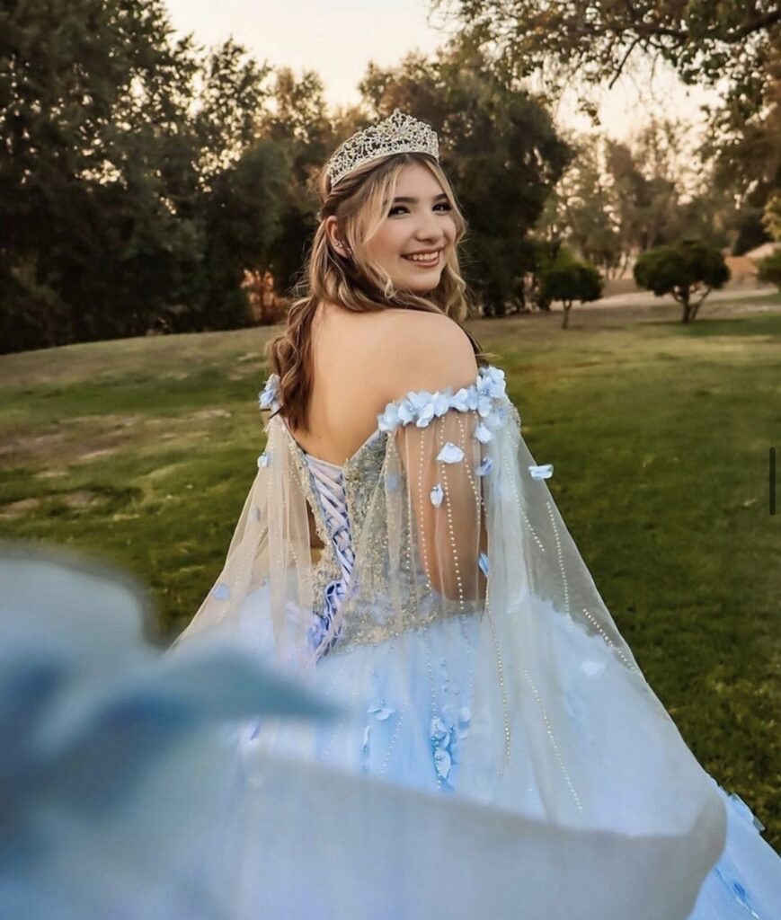 Quinceanera wearing a flowing blue ballgown and tiara looking over her shoulder smiling at the camera.
