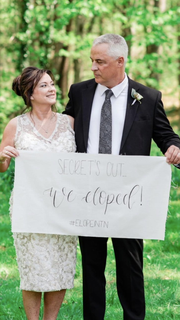 The bride, wearing a knee length lace wedding dress with a v-neckline looks up at the groom. The groom is wearing a black suit with a white shirt and gray tie. They are holding an elopement sign for Elope in Tennessee.