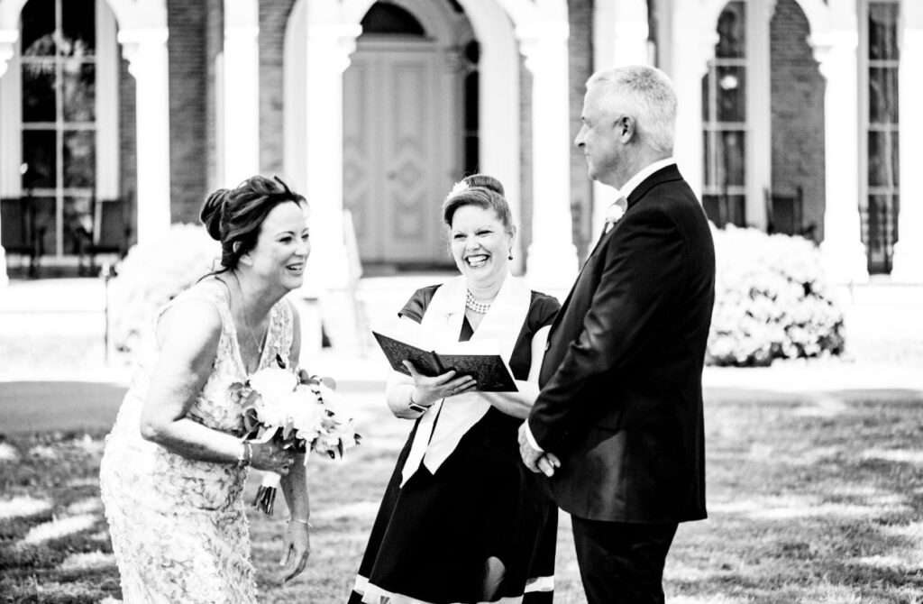 Bride, holding a large bouquet of white flowers is wearing a knee length lace wedding dress. She is laughing at something the groom said. The groom is wearing a black suit. They stand in front of Oaklands Mansion with their officiant who is also laughing. The officiant is wearing a black dress with a white officiant sash. 