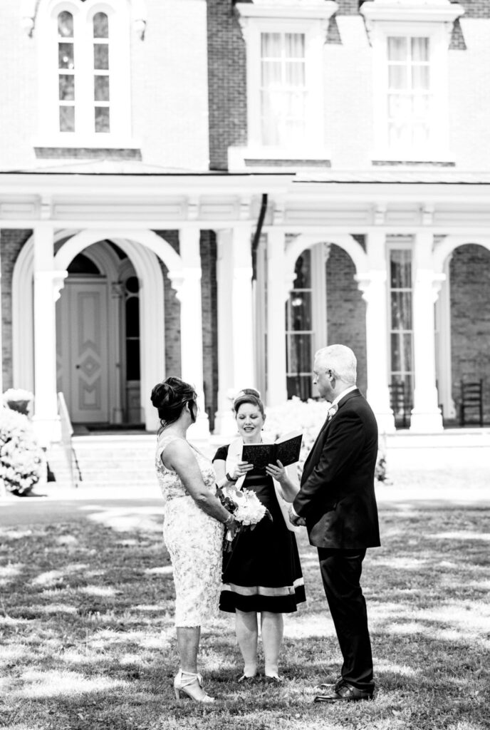 Bride, holding a large bouquet of white flowers is wearing a knee length lace wedding dress. The groom who is wearing a black suit. They stand in front of Oaklands Mansion facing their officiant who is wearing a black dress with a white officiant sash. 