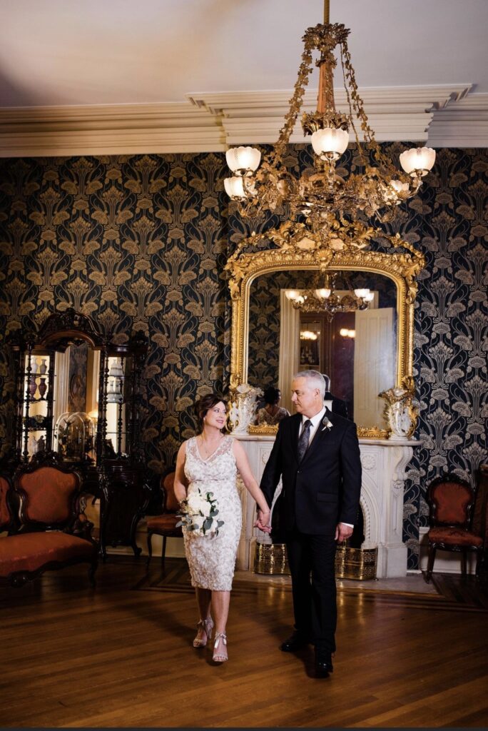 The bride, holding a large white bouquet of flowers wears a knee length v-neck lace wedding dress. She is holding the the groom's hand. He is wearing a black suit with a white shirt and gray tie. They walk in front of an elaborate gold gilded mirror in the front parlor of Oaklands Mansion. The wall paper is dark blue with gold detailing. There is a filigree gas chandelier hanging above their heads. 