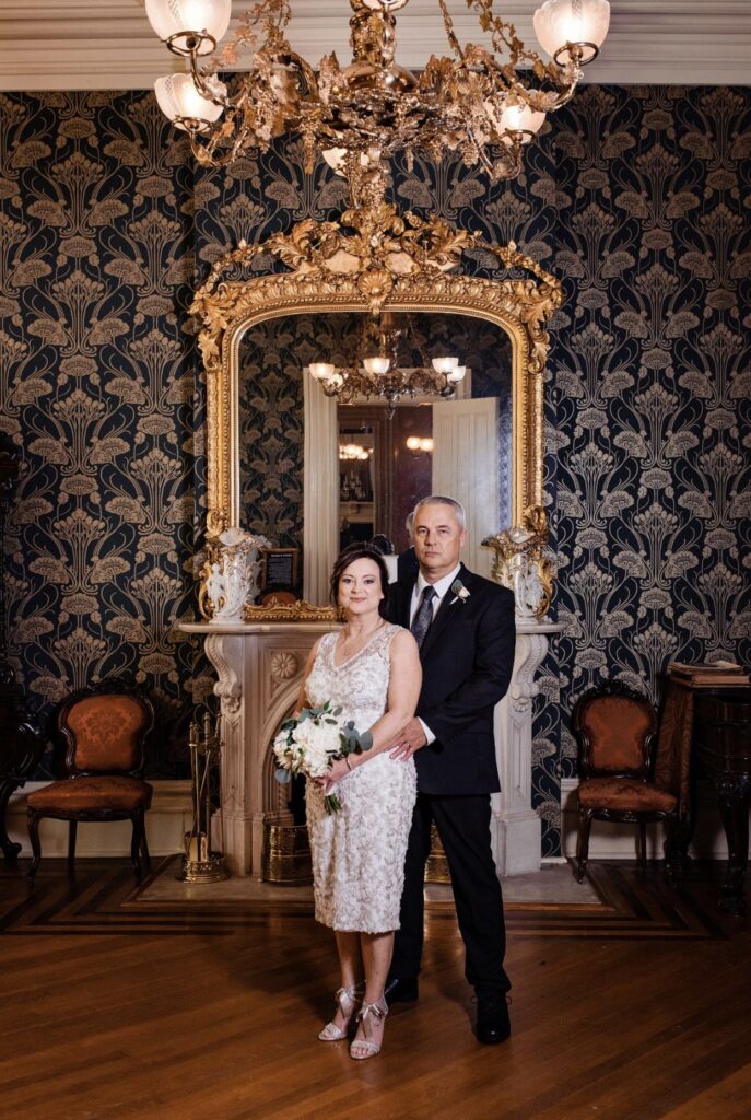 The bride, holding a large white bouquet of flowers wears a knee length v-neck lace wedding dress. She is standing back to front with the groom who is wearing a black suit with a white shirt and gray tie in front of an elaborate gold gilded mirror in the front parlor of Oaklands Mansion. The wall paper is dark blue with gold detailing. There is a filigree gas chandelier hanging above their heads. 