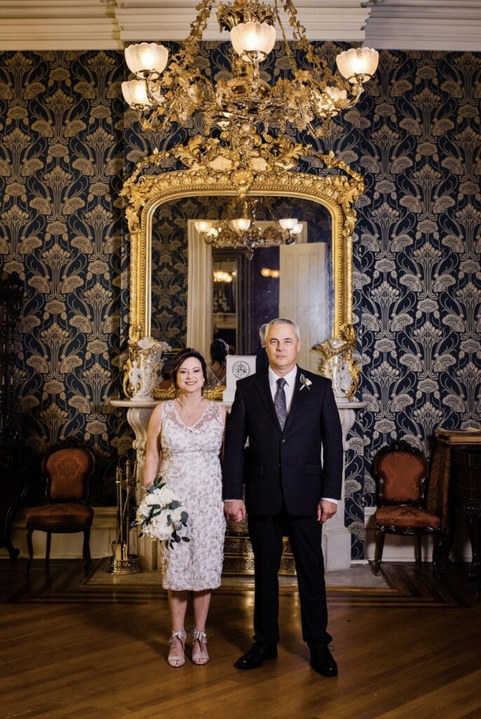 The bride, holding a large white bouquet of flowers wears a knee length v-neck lace wedding dress. She is standing with the groom, holding hands. He is wearing a black suit with a white shirt and gray tie in front of an elaborate gold gilded mirror in the front parlor of Oaklands Mansion. The wall paper is dark blue with gold detailing. There is a filigree gas chandelier hanging above their heads. 