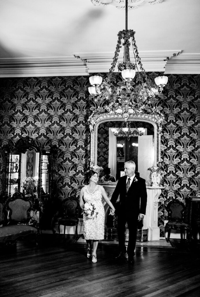 The bride, holding a large white bouquet of flowers wears a knee length v-neck lace wedding dress. She is strolling through the front parlor with the groom. He is wearing a black suit with a white shirt and gray tie in front of an elaborate gold gilded mirror in the front parlor of Oaklands Mansion. The wall paper is dark blue with gold detailing. There is a filigree gas chandelier hanging above their heads. 