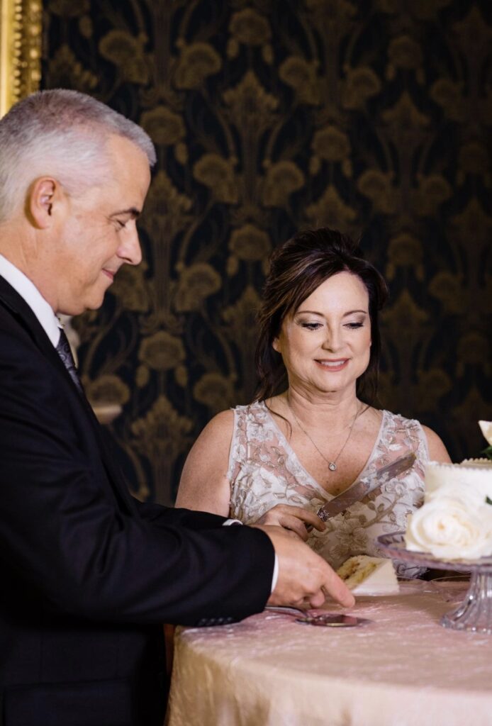 The bride wearing a v-neck lace wedding dress is standing with the groom who is wearing a black suit with a white shirt and gray tie as they cut their small white wedding cake in the front parlor of Oaklands Mansion. The wall paper is dark blue with gold detailing. 