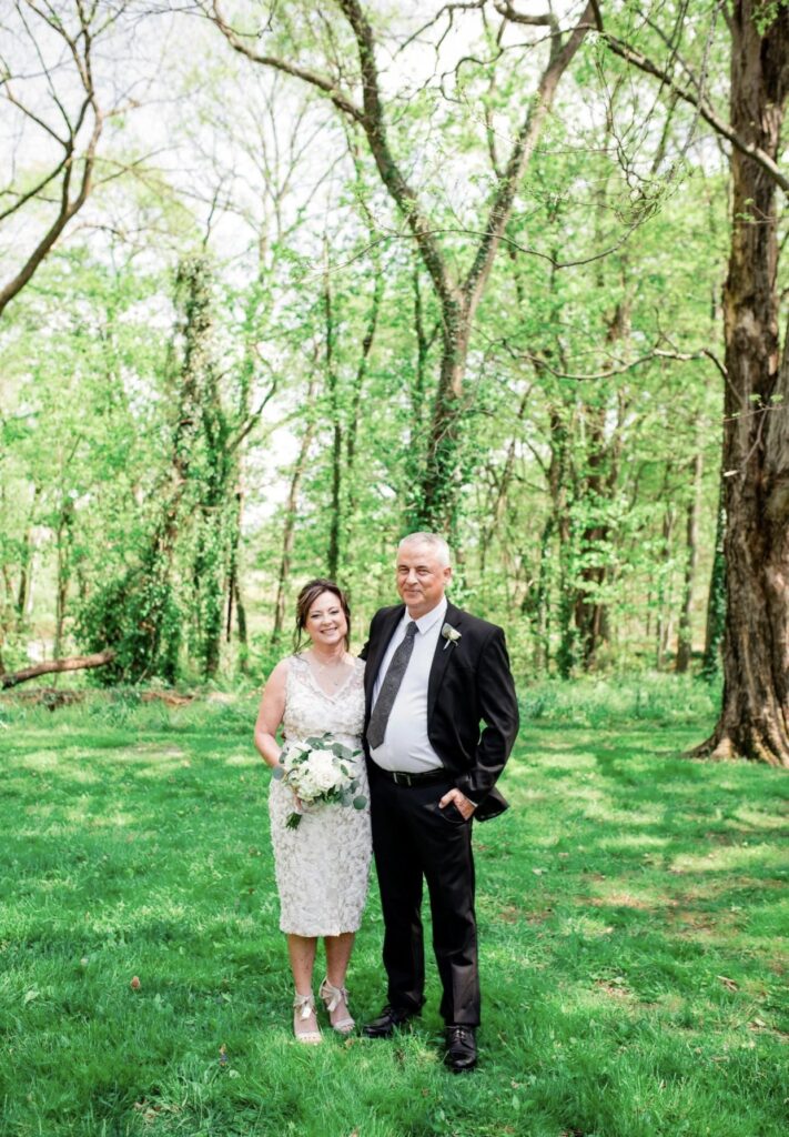 The bride wearing a v-neck lace wedding dress is standing with the groom who is wearing a black suit with a white shirt and gray tie as they smile at the camera. The bride is holding a large white floral bouquet. They are standing in the woods.