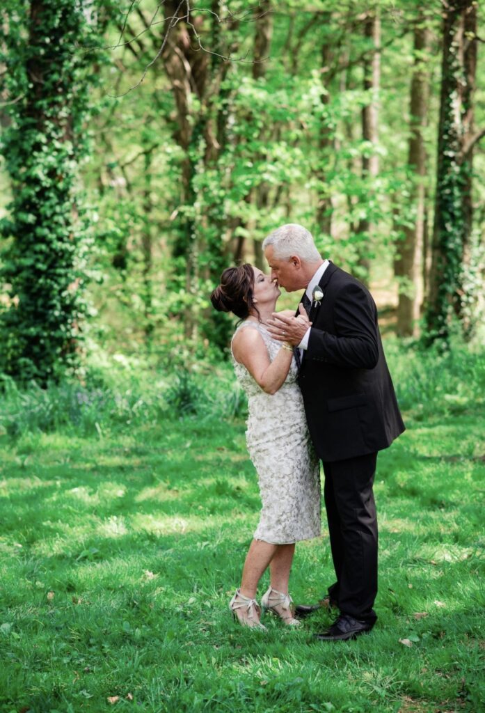 The bride wearing a v-neck lace wedding dress is standing with the groom who is wearing a black suit with a white shirt and gray tie as they embrace and kiss in the woods.