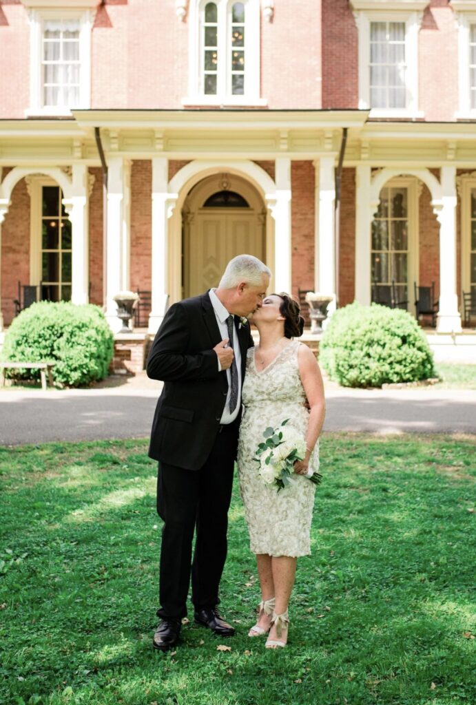 Bride wearing a knee length v-neck lace wedding dress and a groom wearing a black suit with a white shirt and gray tie, kiss in front of Oaklands Mansion