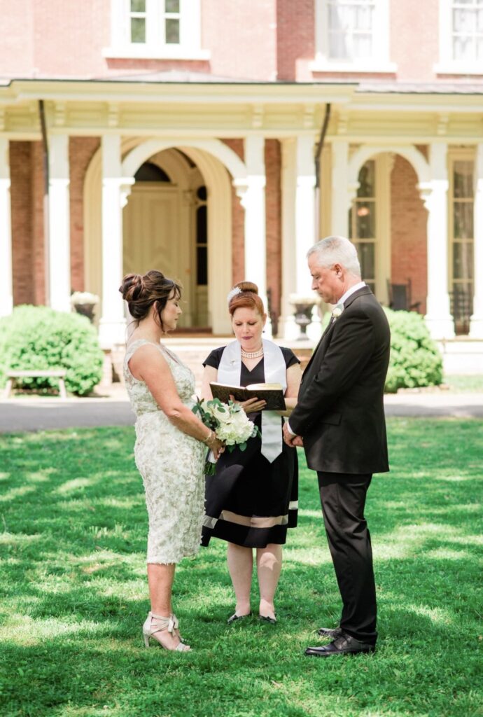 Bride, holding a large bouquet of white flowers is wearing a knee length lace wedding dress. She is holding hands with the groom who is wearing a black suit with a white shirt. They stand in front of Oaklands Mansion with their officiant, from Elope in Tennessee, is wearing a black dress with a white officiant sash. 