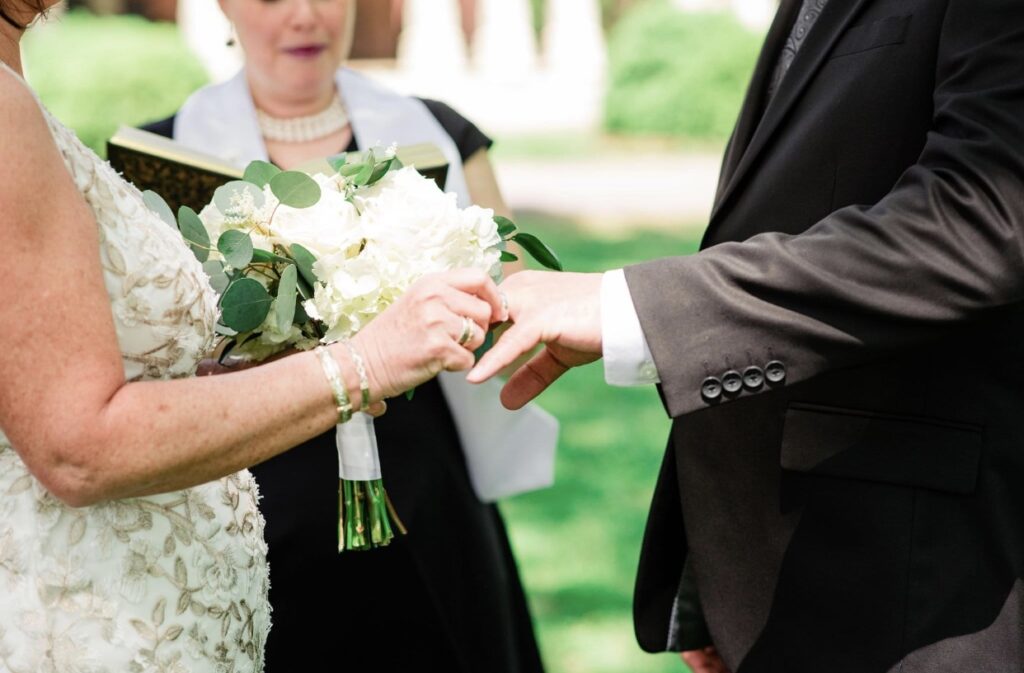 The bride, holding a large bouquet of white hydrangeas and roses puts a wedding band on the groom's finger. 