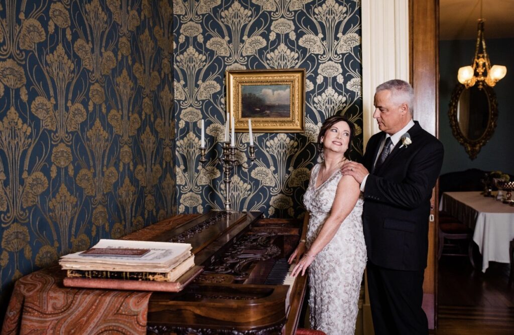 The bride, wearing a v-neck lace wedding dress, is standing with the groom who is wearing a black suit with a white shirt and gray tie in front of an 1850's Chickering piano in the front parlor of Oaklands Mansion. The wall paper is dark blue with gold detailing. 