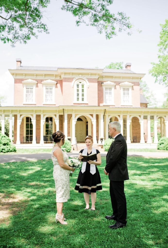 Bride, holding a large bouquet of white flowers is wearing a knee length lace wedding dress. She is holding hands with the groom who is wearing a black suit with a white shirt. Their officiant who is wearing a black dress with a white officiant sash and is reading from a book. They are standing in front of Oaklands Mansion.