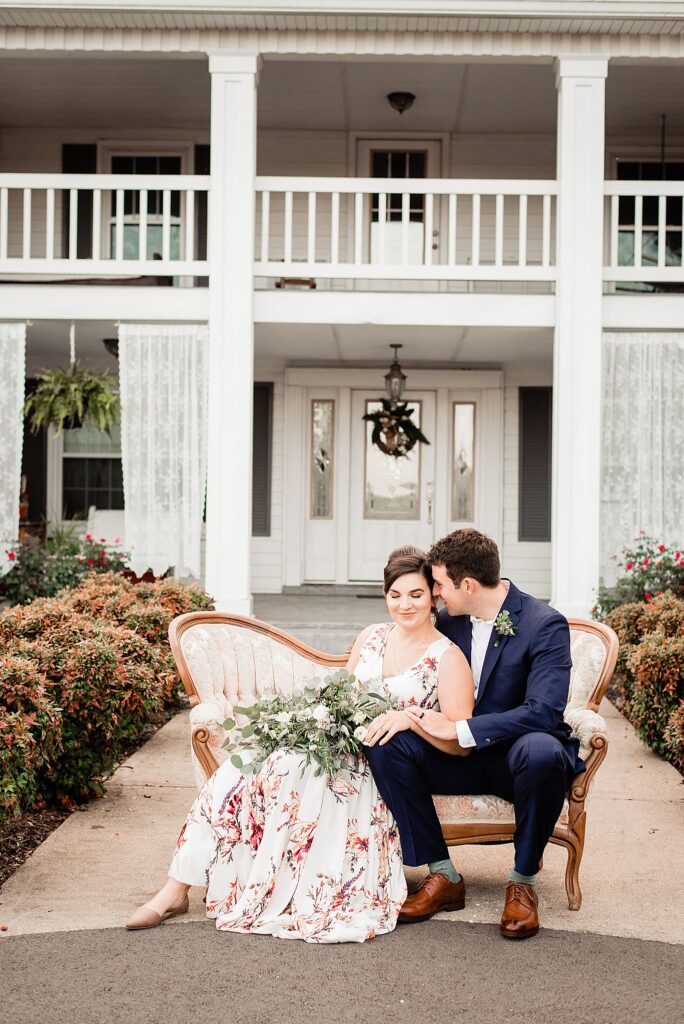 Bride in a flowing floral gown and groom in a navy suit embrace on an ivory brocade sofa in front of an elegant white farmhouse in Nashville, Tennessee