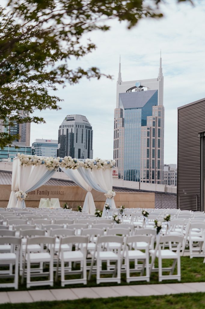A view of downtown Nashville from a rooftop wedding ceremony at The Country Music Hall of Fame. The draped arbor is blue and white with clusters of white florals 