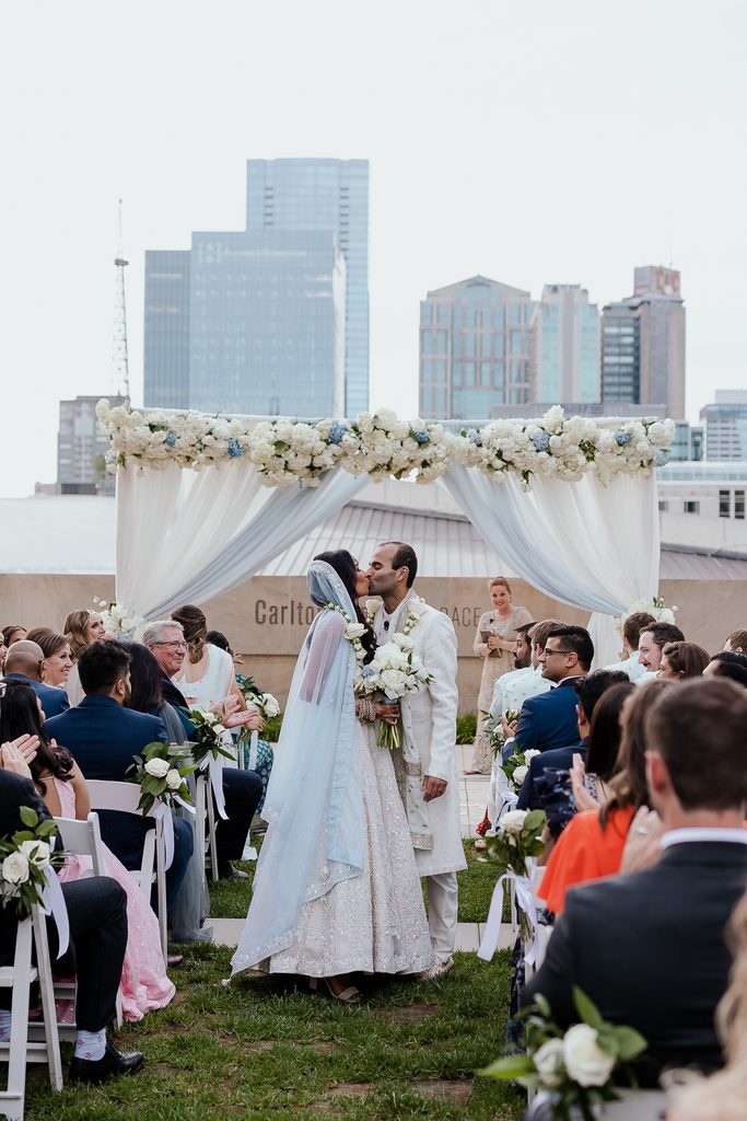 Bride and groom kissing on the  rooftop of The Country Music Hall of Fame during their wedding ceremony. The draped arbor is blue and white with clusters of white florals 