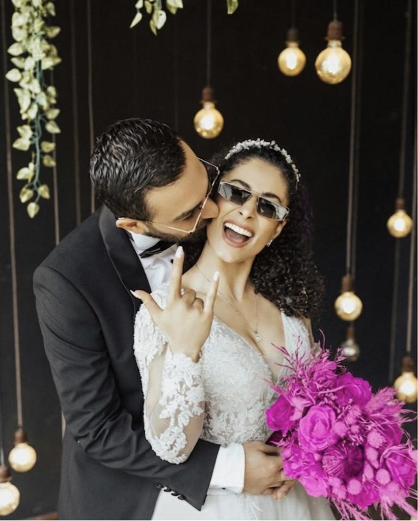 Bride and groom wearing sun glasses laugh and embrace. The bride is hold ing a hot pink monochromatic bouquet and is wearing a long sleeved lace gown. The groom kisses her cheek. He is wearing a black tuxedo. There is a hanging Edison bulbs chandelier behind them. 