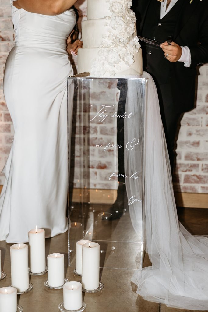 The bride and groom stand on their side of their white wedding cake with white sugar flowers and a long tulle veil sits on a clear acrylic pillar next to a cluster of white pillar candles at Steel Magnolia Barn